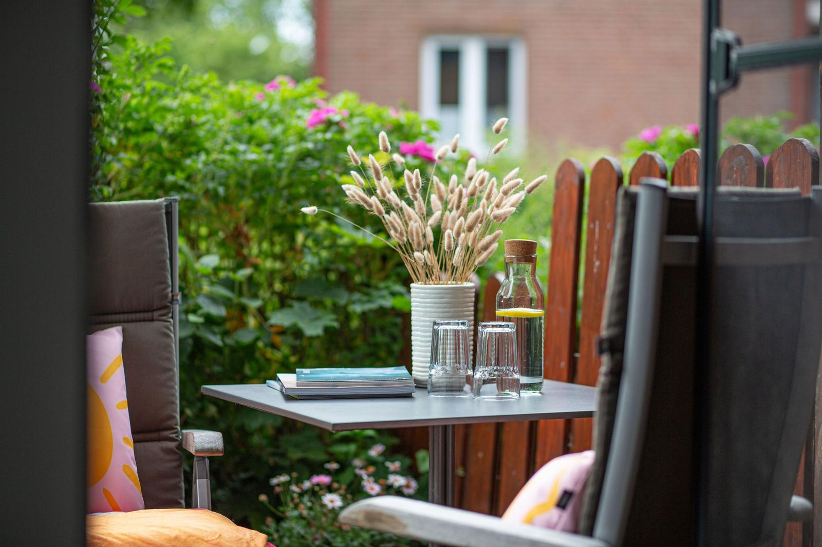 Terrace with table, chairs, vases, and plants. Background with hedge and house.