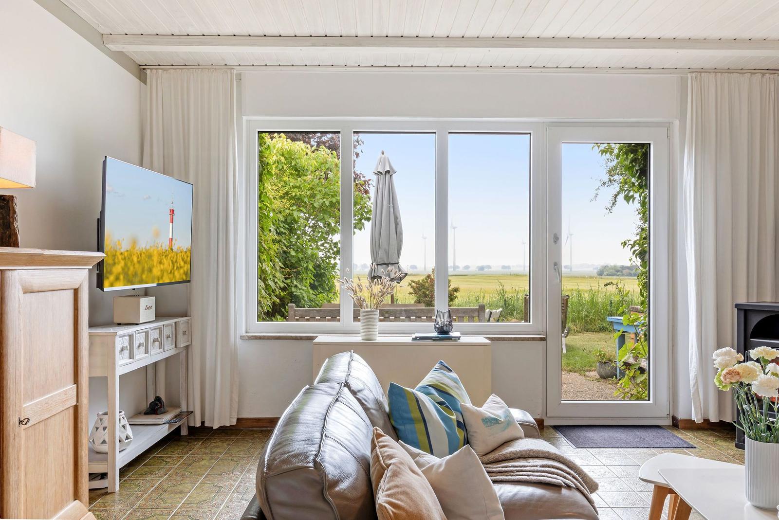 Wohnzimmer mit Sofa, Fernseher und Blick auf Garten und Felder.