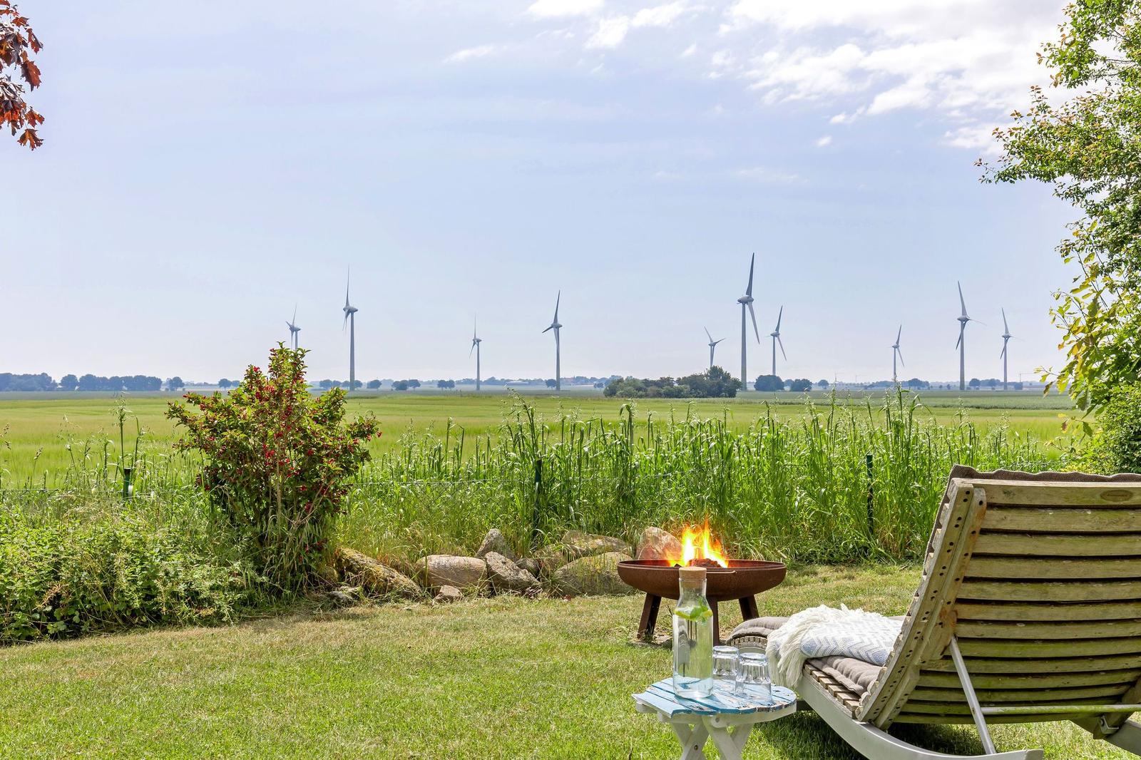 Außenbereich mit Liegestuhl, Feuerkorb und Blick auf Windräder im Grasland.