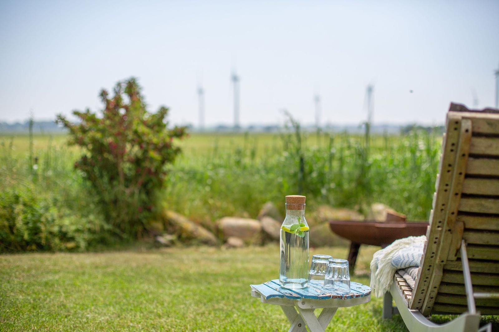 Außenbereich mit Liegestuhl, Tisch und Wasserflasche. Im Hintergrund sind Windräder und Grünfläche zu sehen.