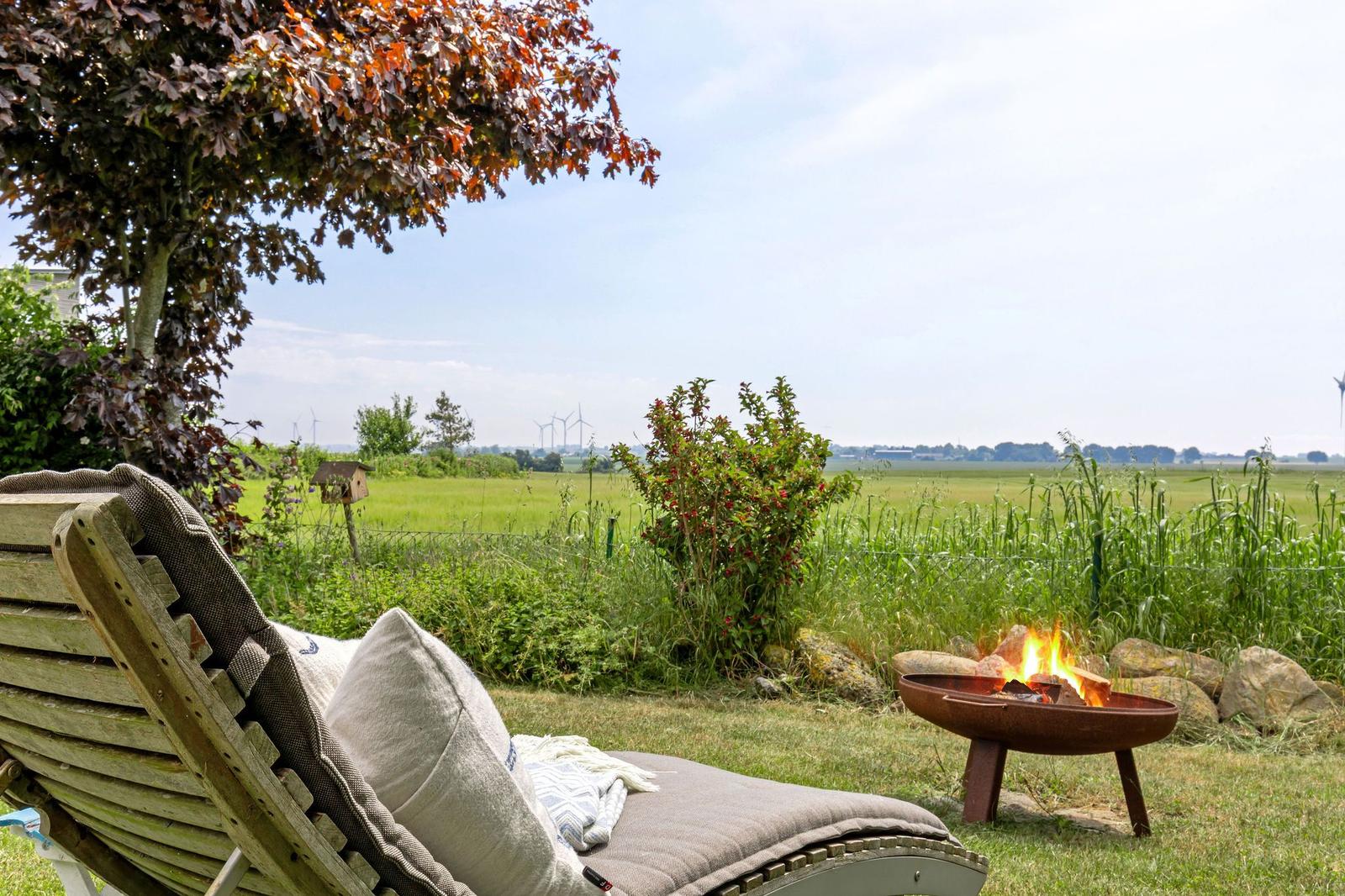 Holzliegestuhl mit Kissen neben brennendem Feuerkorb im Garten mit Blick auf Felder und Windräder.