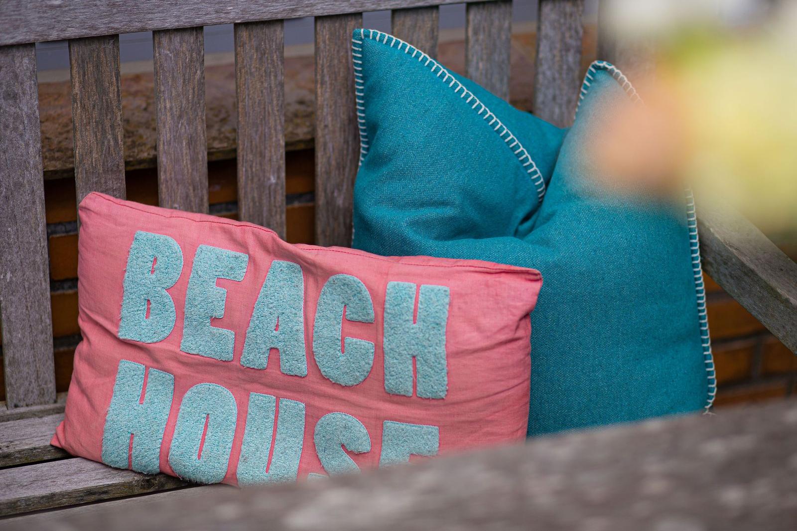 Auf einer Holzbank liegen zwei Kissen, eines mit der Aufschrift BEACH HOUSE.