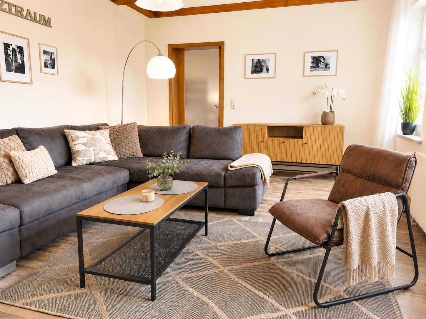 Wohnzimmer mit grauem Sofa, braunem Sessel und Holztisch auf Teppich.