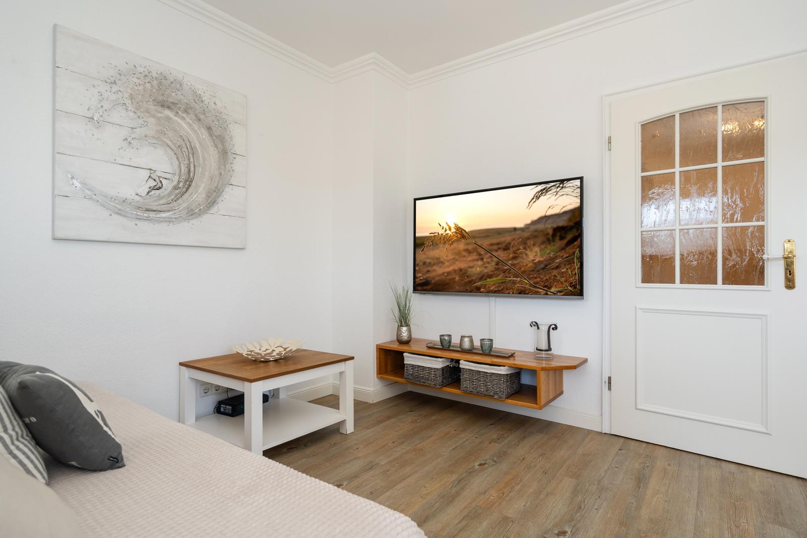 Wohnzimmer mit Fernseher, Tisch und Wandbild. Holzfußboden und weiße Wände.