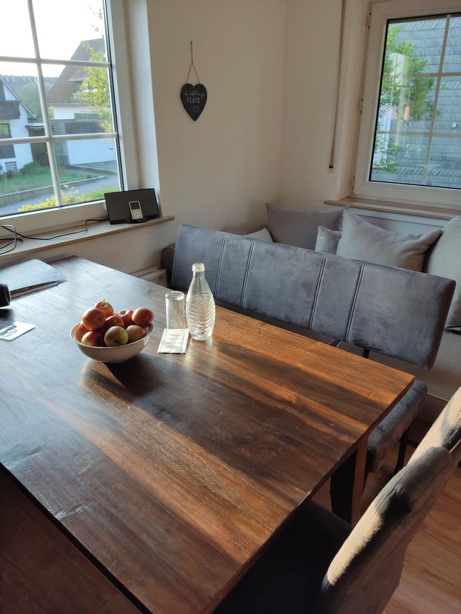 Holztisch mit Obstschale und Wasserflasche, graue Sitzbank und Fenster mit Blick.