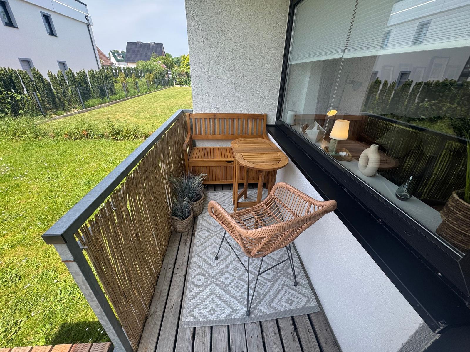 Terrasse mit Holzbank, Tisch und Rattanstuhl. Grünfläche im Hintergrund.