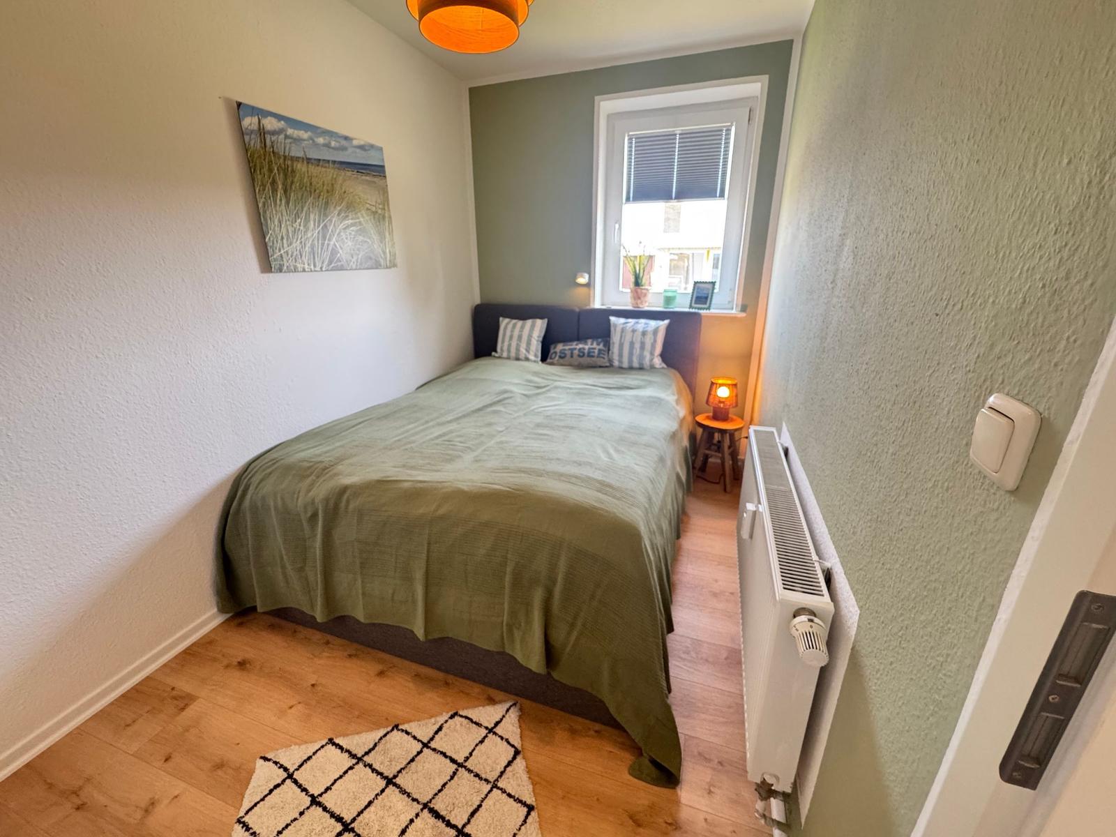 Schlafzimmer mit Bett, Fenster und Heizung. Wandbild und Teppich sind sichtbar.