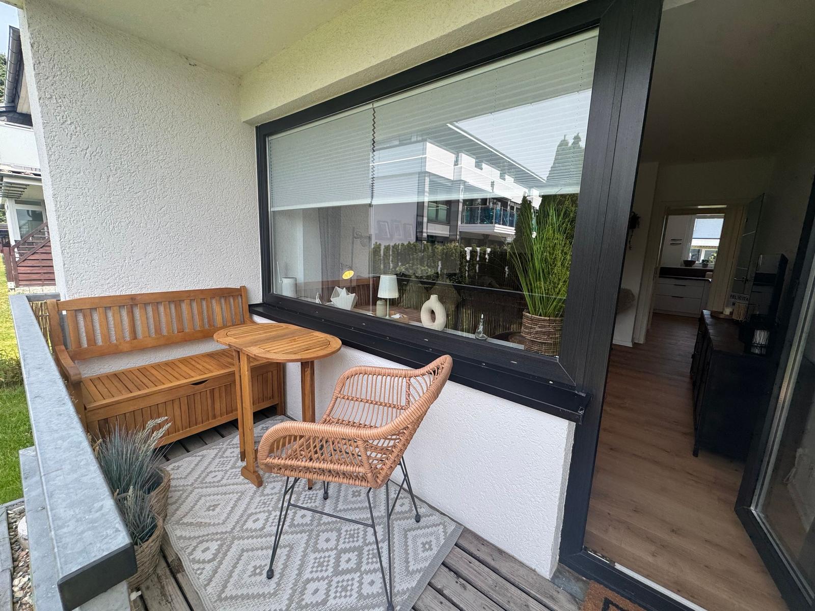 Terrasse mit Holzbank, Tisch und Rattan-Stuhl. Große Fensterfront zum Innenraum.