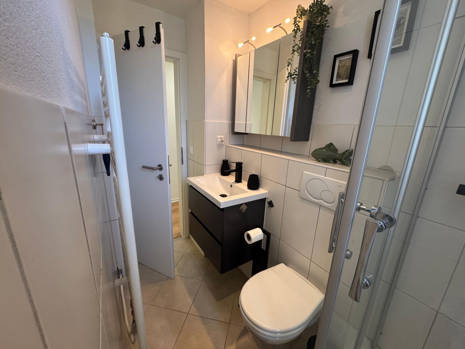Badezimmer mit Dusche, Waschbecken und Toilette. Spiegel und Pflanze an der Wand.