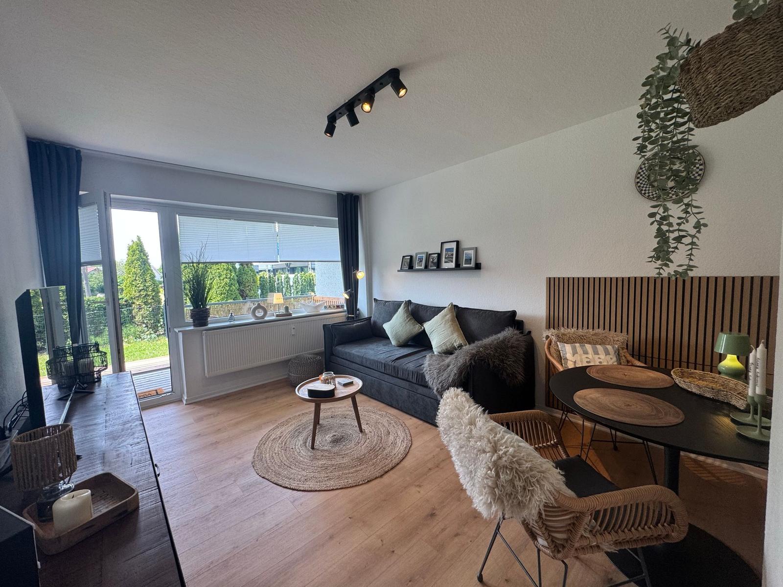 Wohnbereich mit Sofa, Esstisch und Balkontür. Holzboden und Pflanzen.