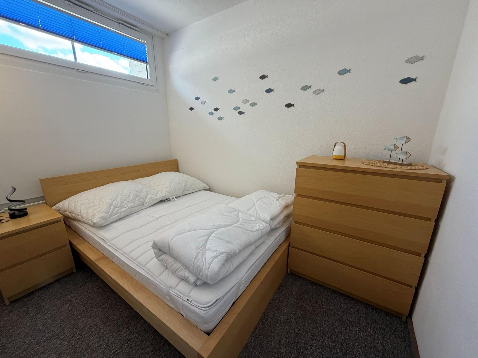 Schlafzimmer mit Bett, Kleiderschrank und Fenster mit blauen Jalousien.