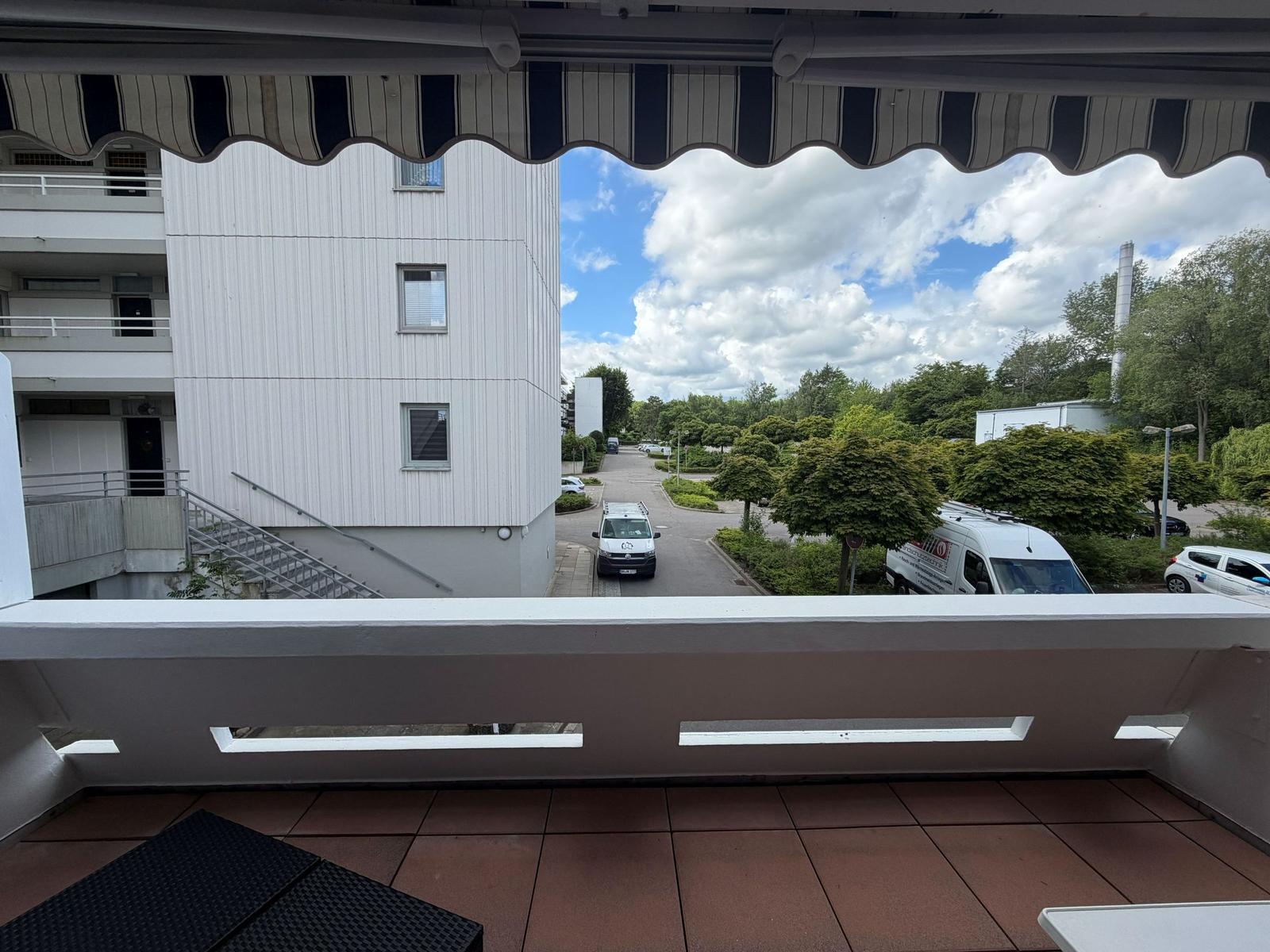 Balkon mit Blick auf Wohngebäude und grüne Umgebung unter blauem Himmel mit Wolken.