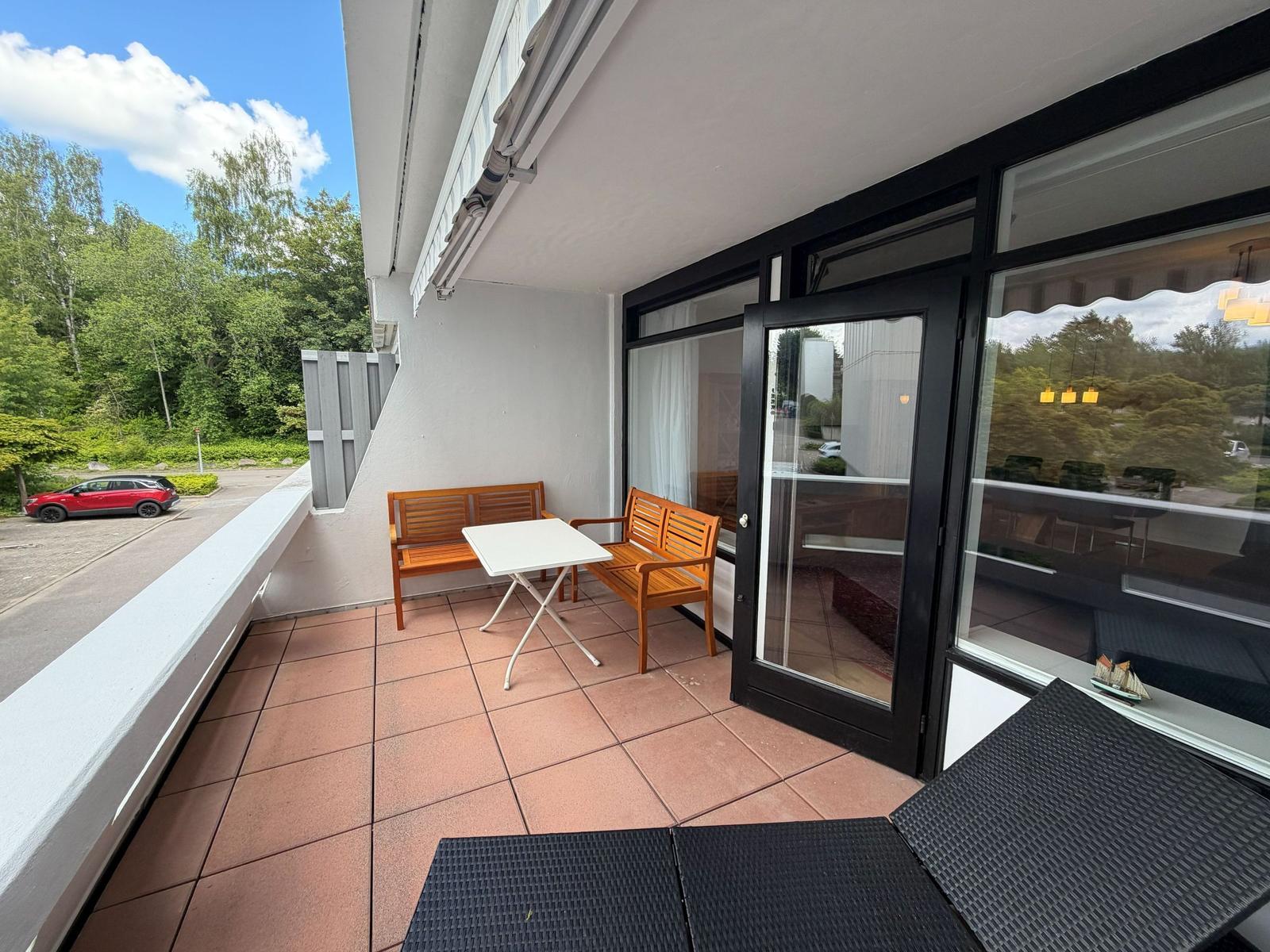 Terrasse mit Tisch, zwei Bänken und Liegeflächen. Blick auf grüne Umgebung und Parkplatz.