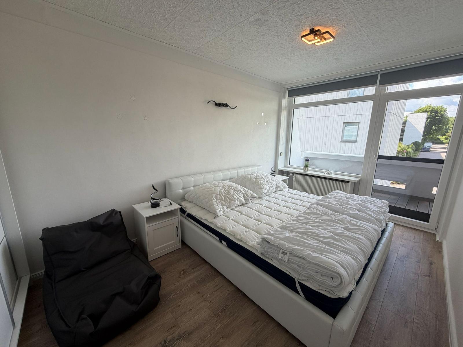 Schlafzimmer mit Bett, Nachttisch und Sitzgelegenheit. Große Fensterfront mit Blick auf Balkon.