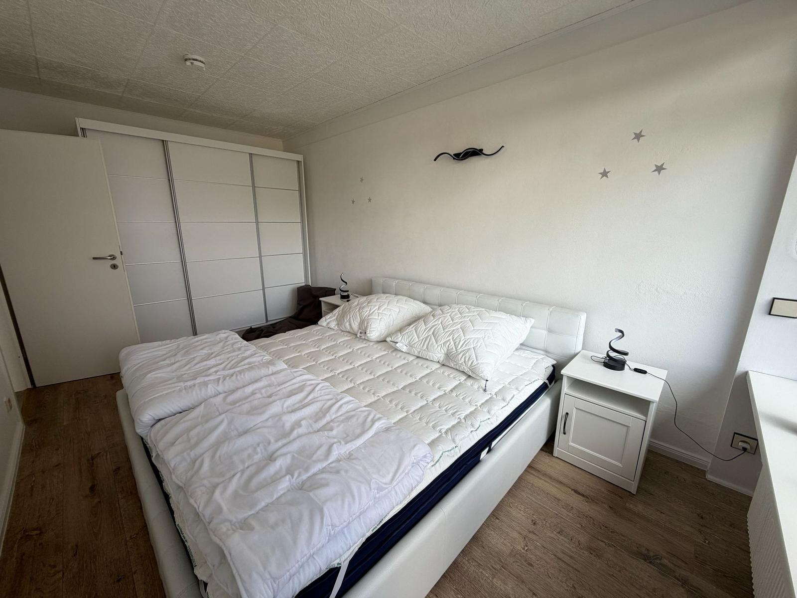 Schlafzimmer mit weißem Bett, Nachttisch und Schrank. Holzfußboden und Wanddekoration.