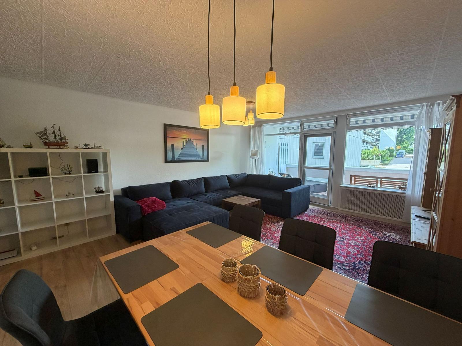 Wohn- und Essbereich mit Sofa, Tisch und Hängelampen. Fenster mit Blick nach draußen.