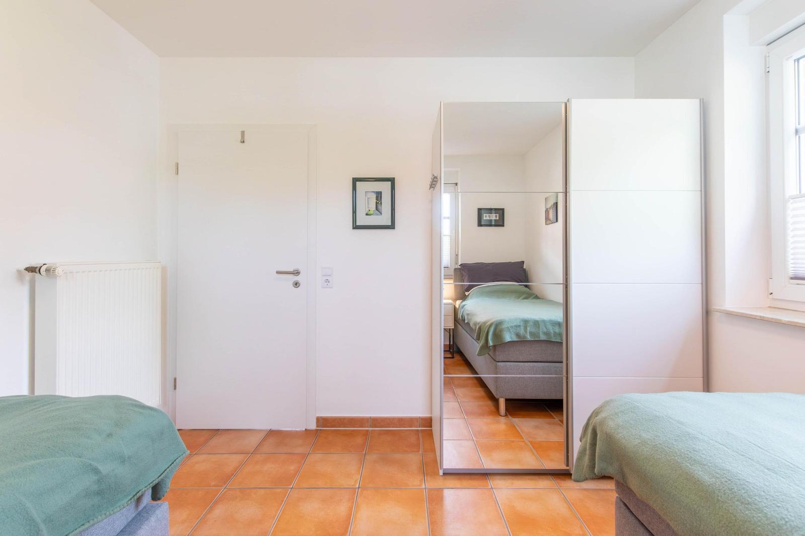 Room with two beds, mirrored wall, and window. Tile floor and white walls.