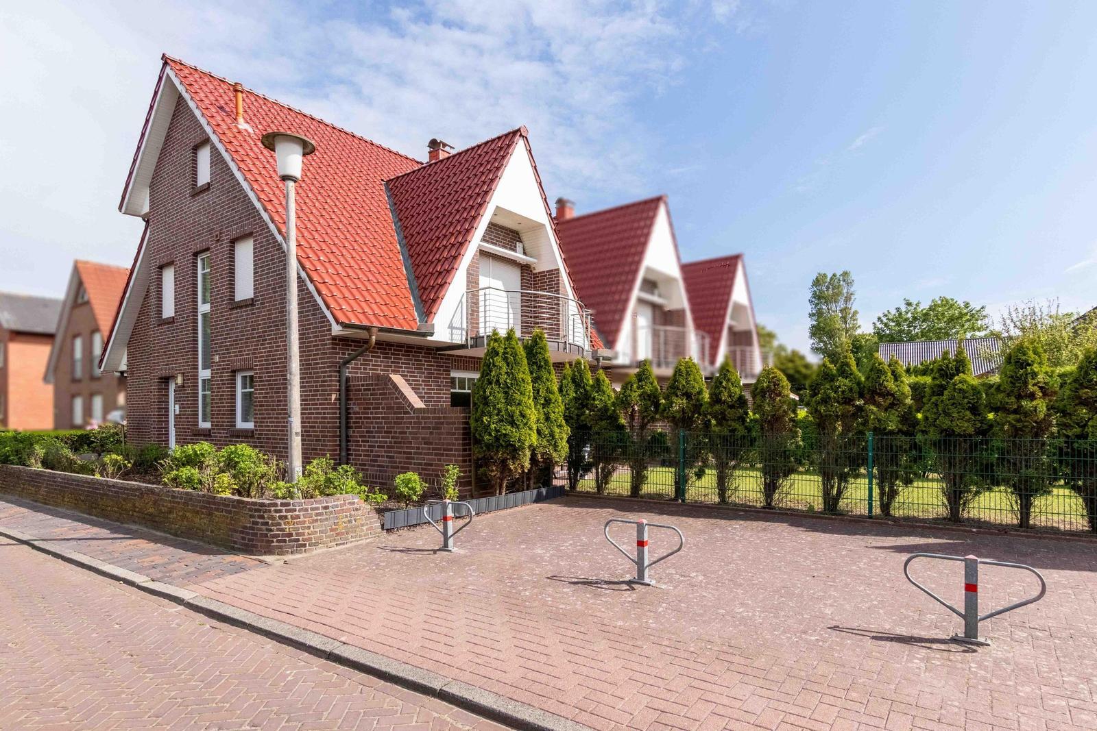 Red tiled roof, brown facade, paved street with bike racks and green hedge.