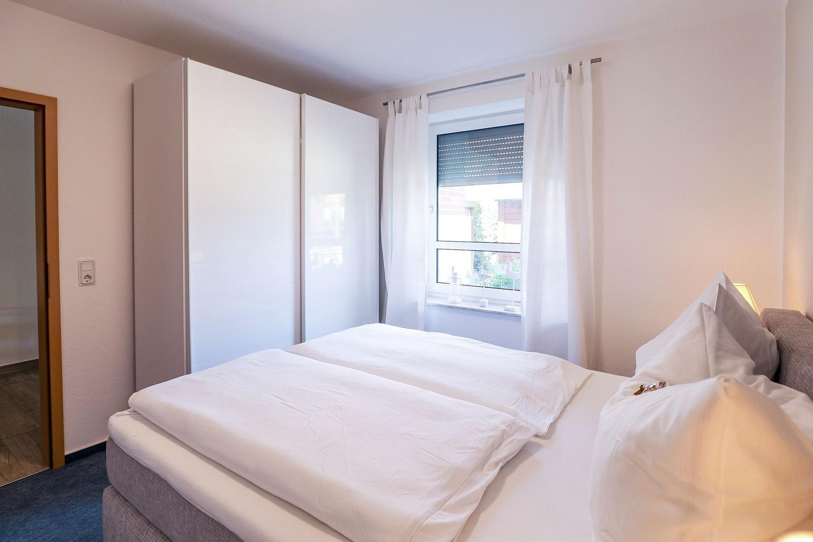 Schlafzimmer mit Doppelbett, weißen Vorhängen und Schrank. Fenster mit Rollladen.