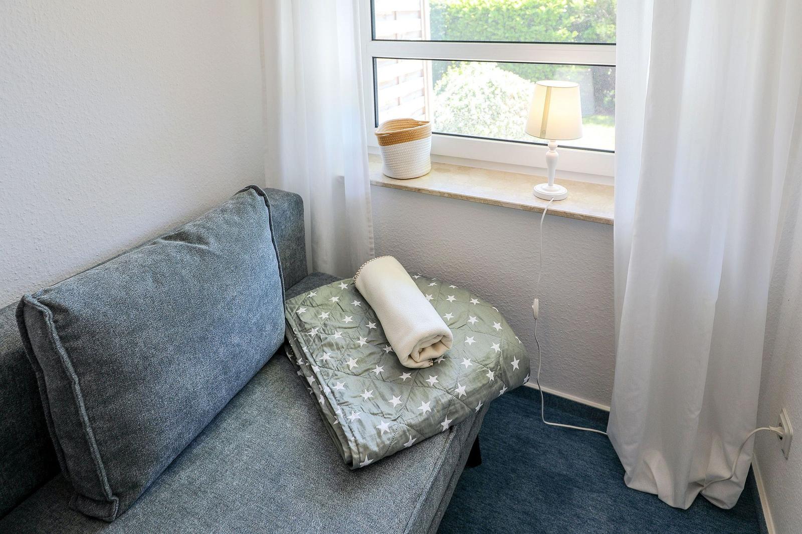 Ein grauer Sessel mit Decke und Kissen neben einem Fenster mit Lampe und Vorhang.