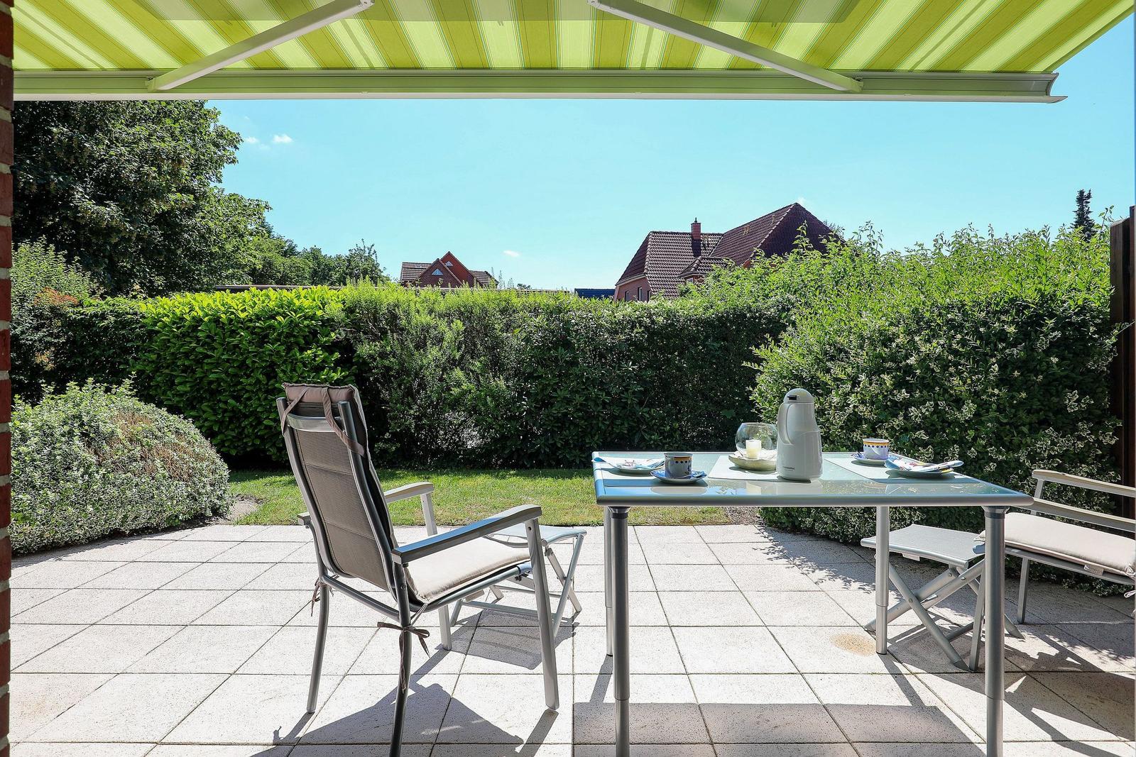 Terrasse mit Tisch, Stühlen und Sonnenschutz. Blick auf grünen Zaun und Häuser.