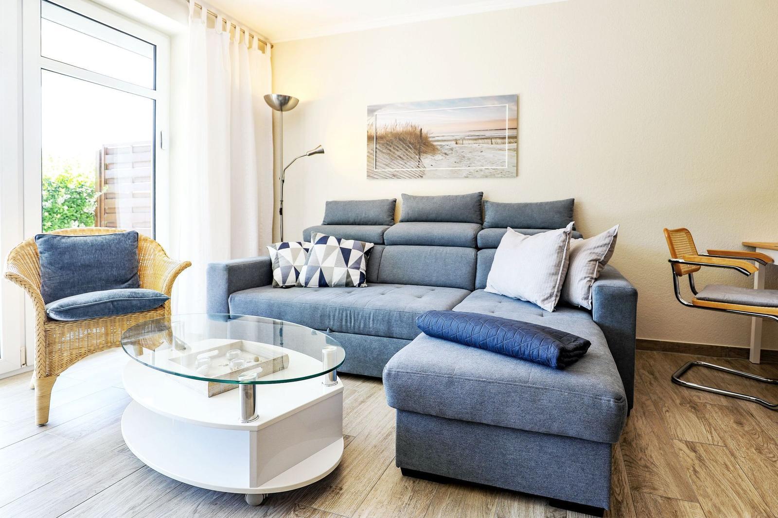 Ein Wohnzimmer mit grauem Sofa, Glas-Tisch und Wicker-Stuhl. Wandbild zeigt Strand.