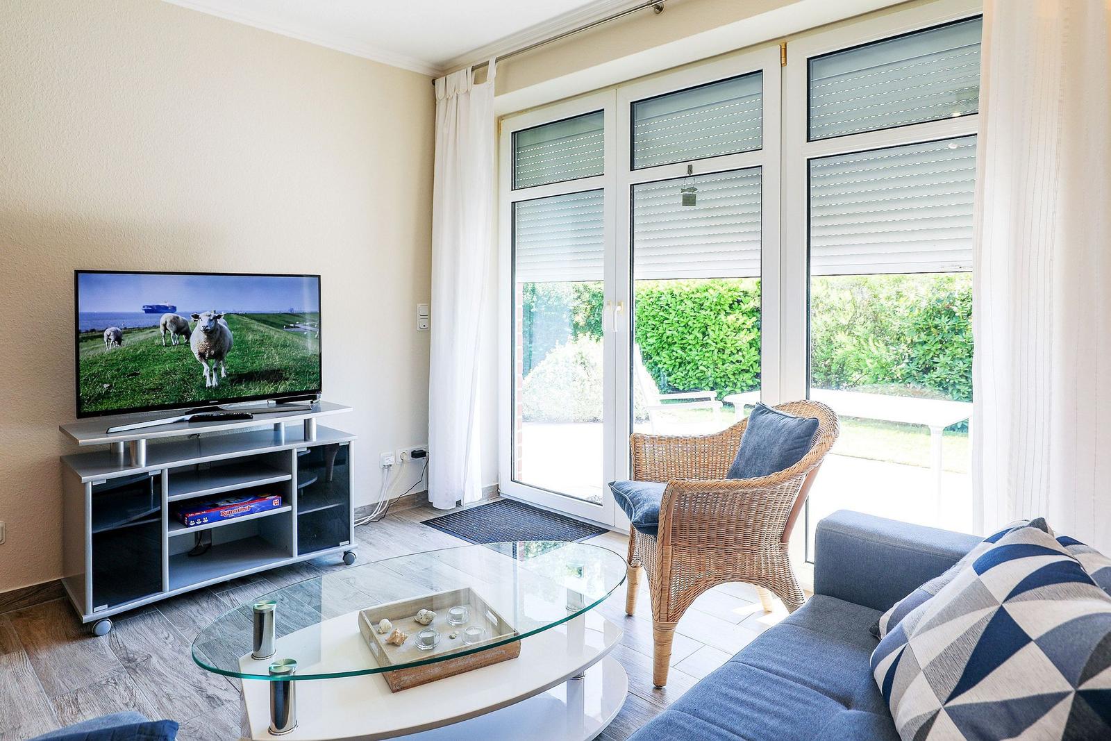 Wohnzimmer mit Sofa, Glas-Tisch und Fernseher. Durchgang zur Terrasse mit Garten.