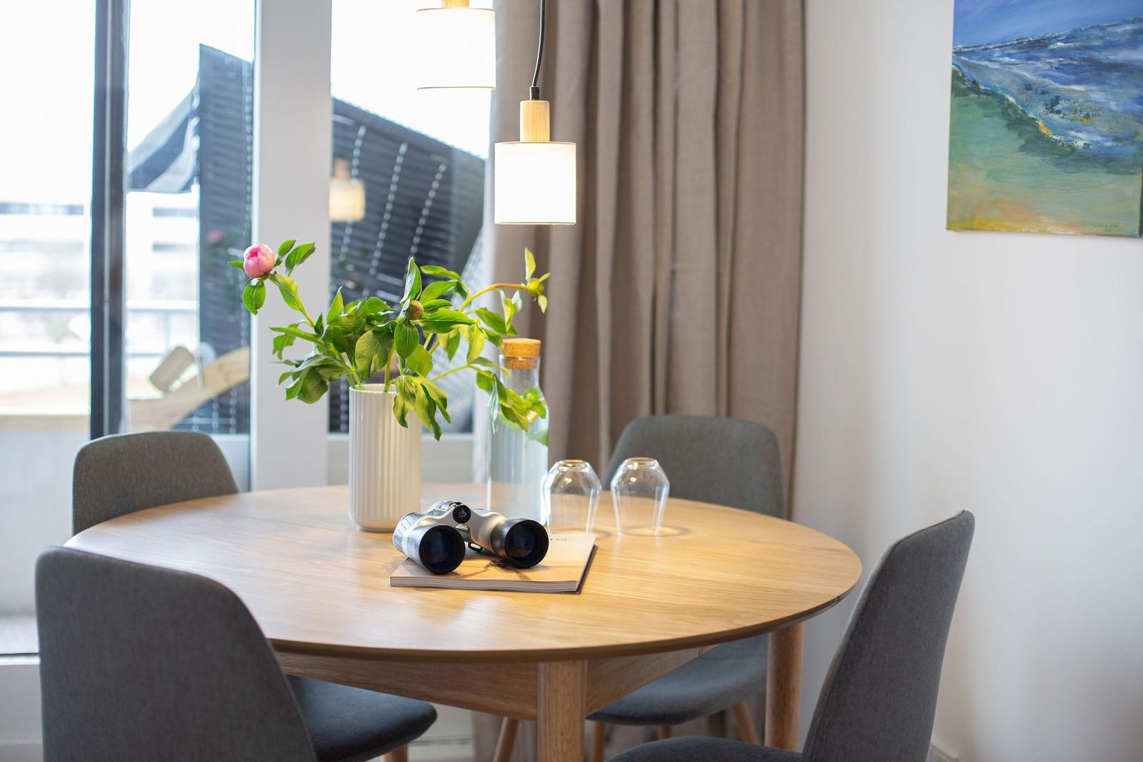Runder Tisch mit Stühlen, Blumen und Fernglas. Fensterblick und Wandbild.