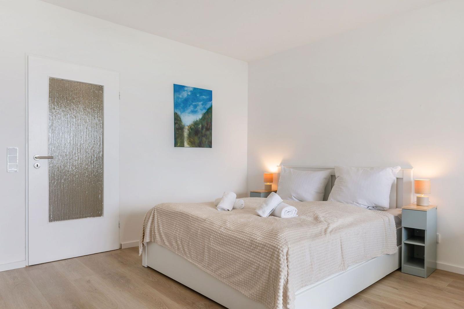 Schlafzimmer mit Bett, Nachttischen und Wandbild. Tür und Holzboden sind sichtbar.