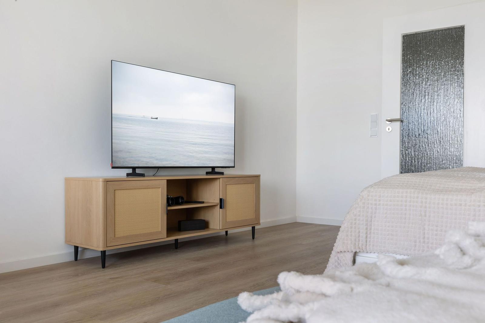 Schlafzimmer mit TV auf Holzregal und Bett mit weißer Bettwäsche.