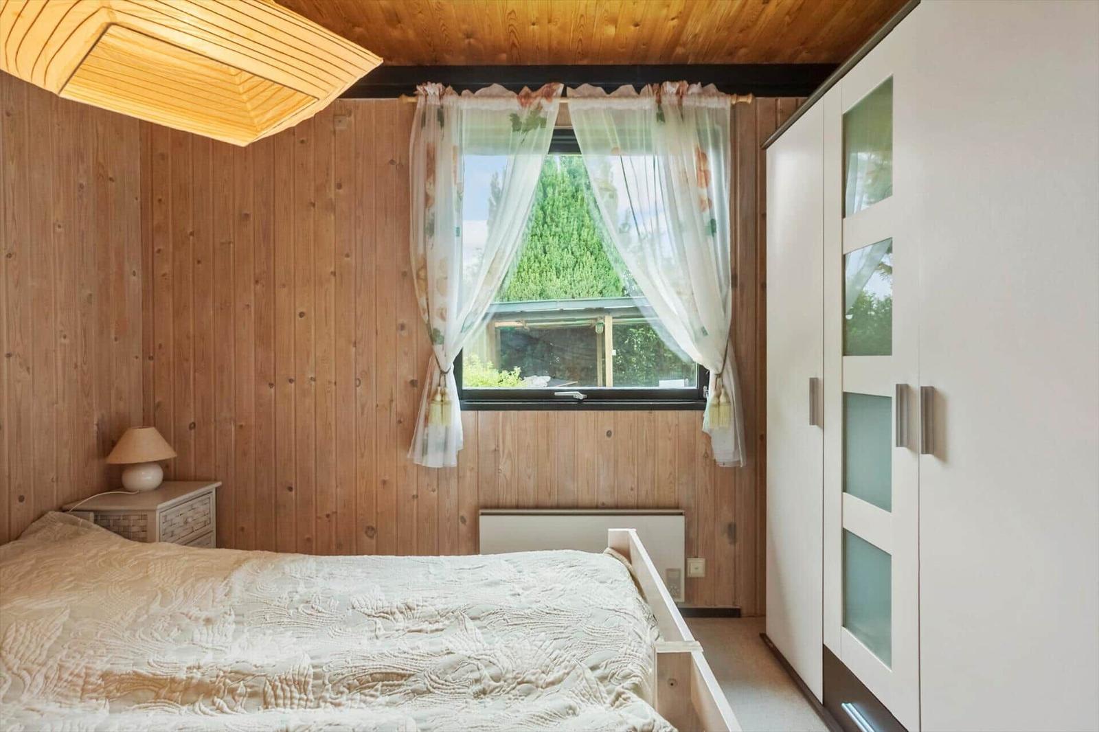 Schlafzimmer mit Holzwand, Bett, Kleiderschrank und Fenster mit Vorhängen.