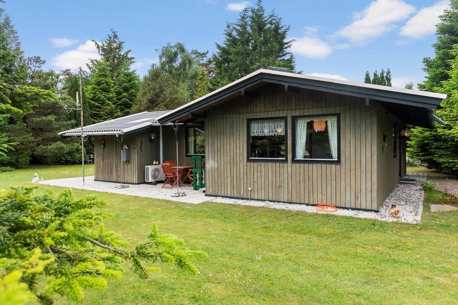 Holzhaus mit Terrasse, Garten und Bäumen. Fenster mit Vorhängen. Außentisch und Stühle.