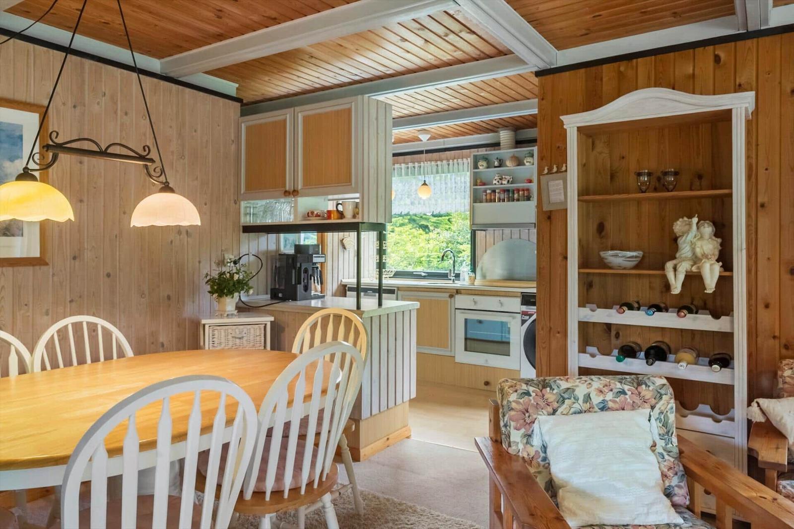 Essbereich mit Holztisch und Stühlen, Küchenbereich mit Fenster, Weinregal und Sessel mit floraler Polsterung.