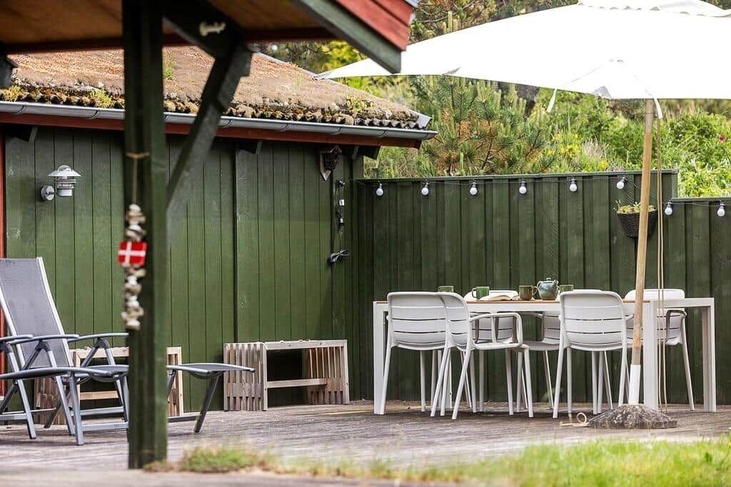 Terrasse mit Tisch, Stühlen und Sonnenschirm. Hintergrund: grüne Holzwand und Garten.