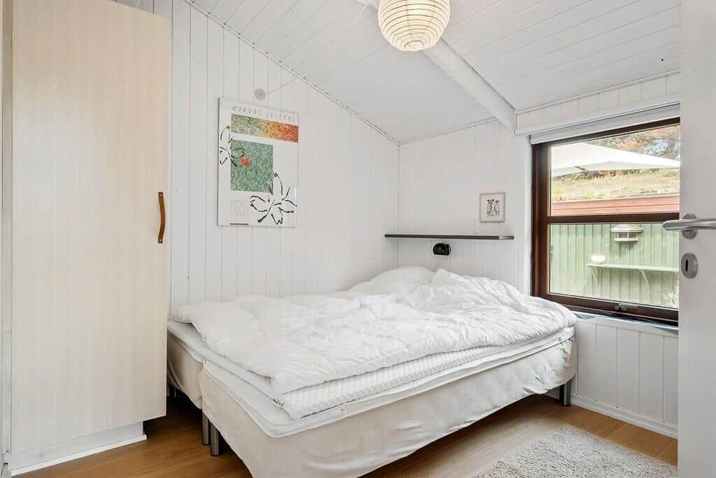 Schlafzimmer mit weißem Bett, Holzwand und Fensterblick auf Garten.