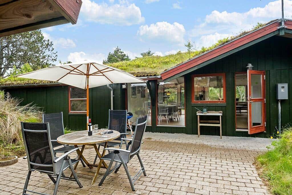 Terrasse mit Tisch, Stühlen und Sonnenschirm vor einem grünen Haus mit Dachgarten.