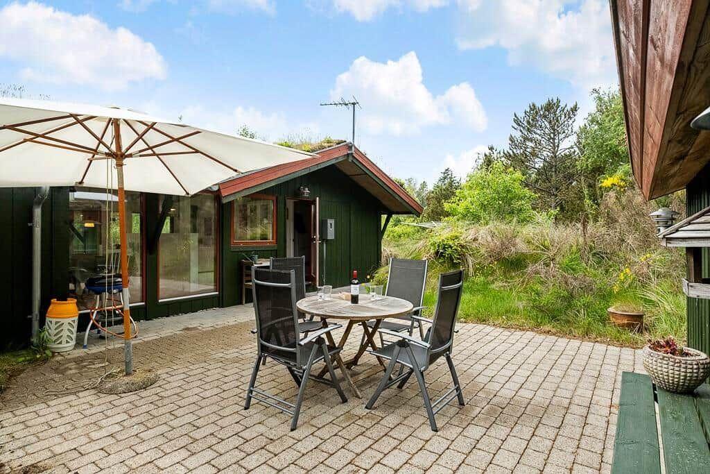 Terrasse mit Tisch, Stühlen und Sonnenschirm vor einem grünen Haus.