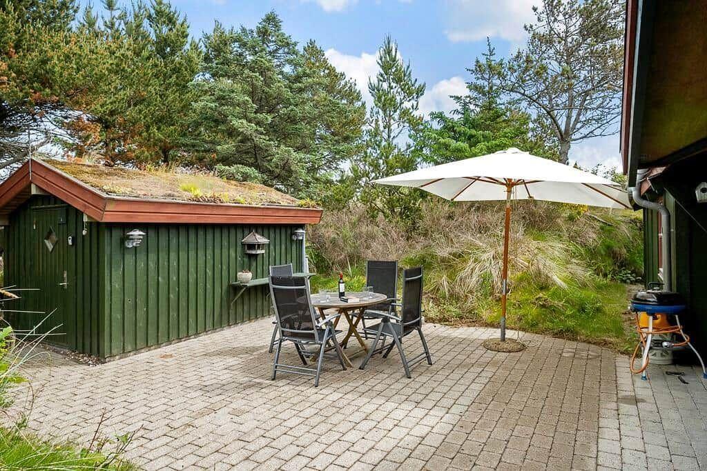 Terrasse mit Tisch, Stühlen und Sonnenschirm neben grünem Holzhaus und Wald.