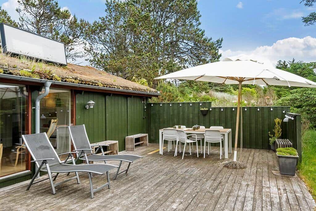 Terrasse mit Sonnenschirm, Tisch, Stühlen und Liegestühlen. Hintergrund: grüne Wand und Bäume.