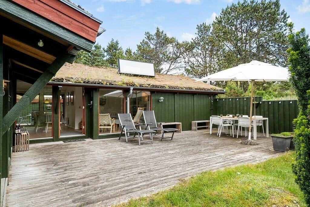 Haus mit Holzterrasse, Gartenmöbel und Sonnenschirm. Grün getönter Holzaufbau mit Glasfront.