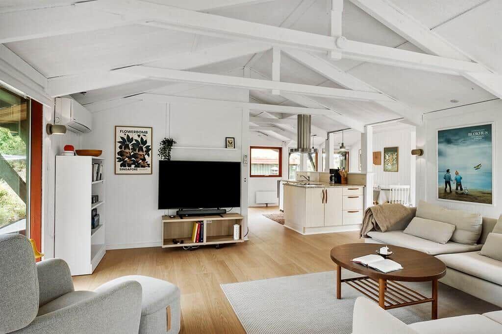 Wohnbereich mit Sofa, Fernseher und Küchenbereich. Holzboden und weiße Wände.