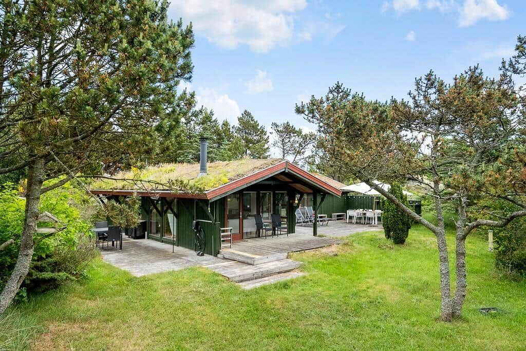 Grüne Hütte mit Terrasse, umgeben von Bäumen und Gras.