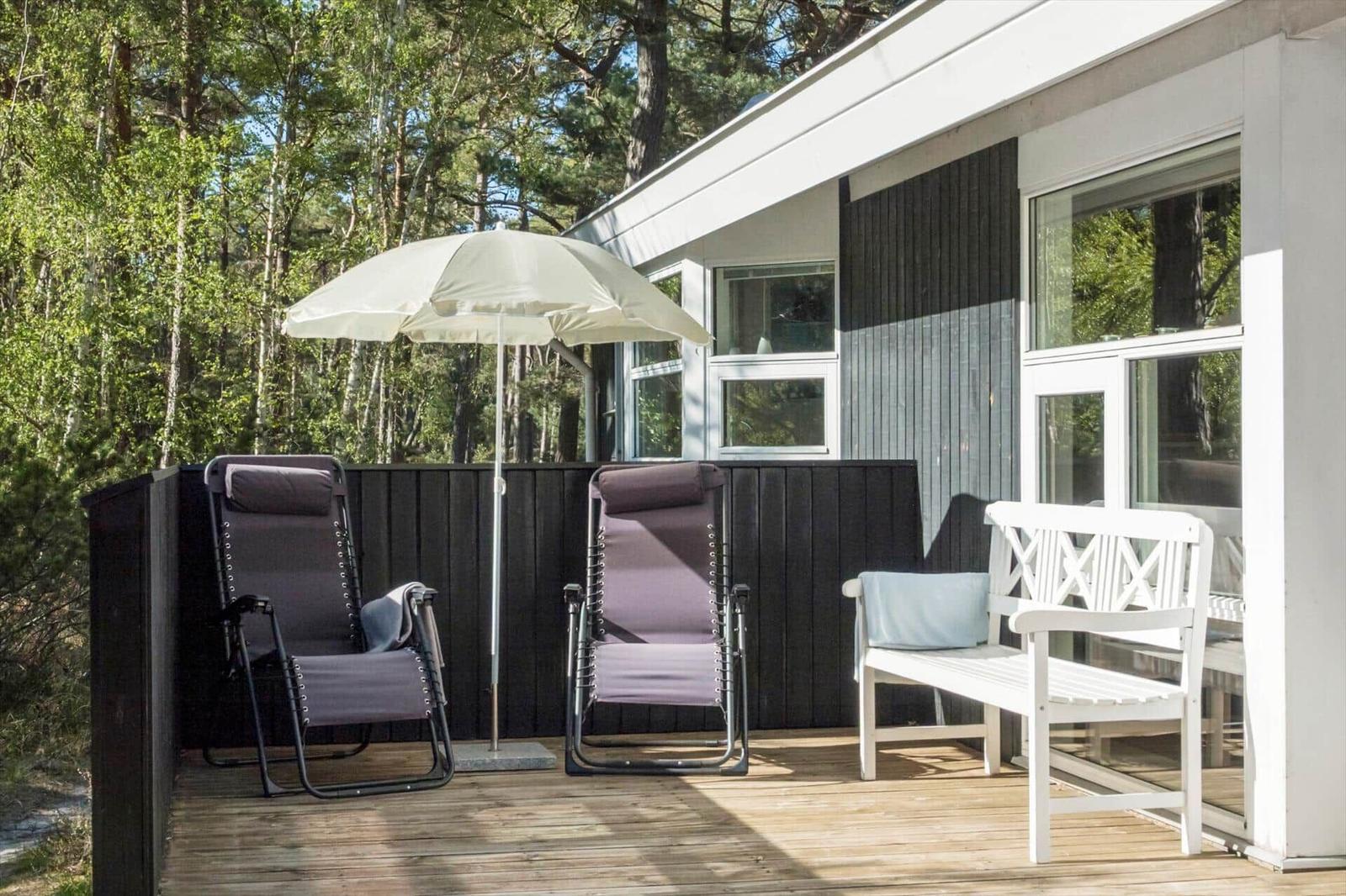 Terrasse mit zwei Liegestühlen, weißer Bank und Sonnenschirm. Hintergrund: Wald und Hauswand.
