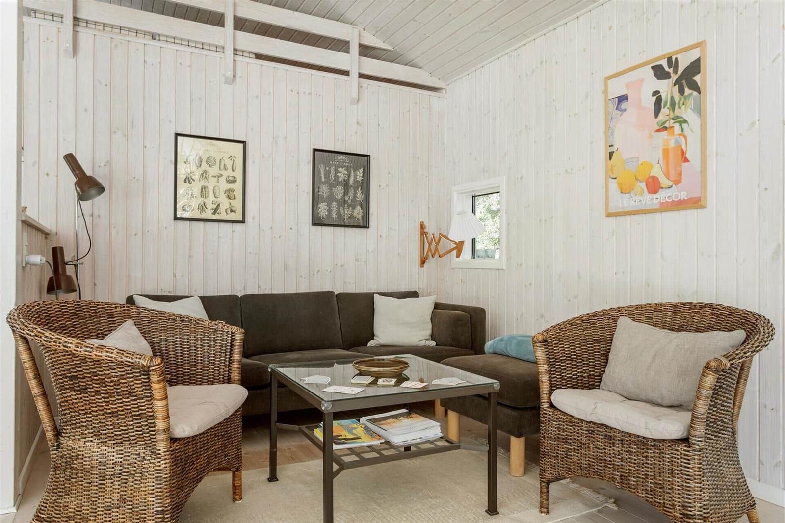 Wohnzimmer mit Sofa, zwei Rattan-Stühlen und Glas-Tisch. Holzwände und Wandbilder.