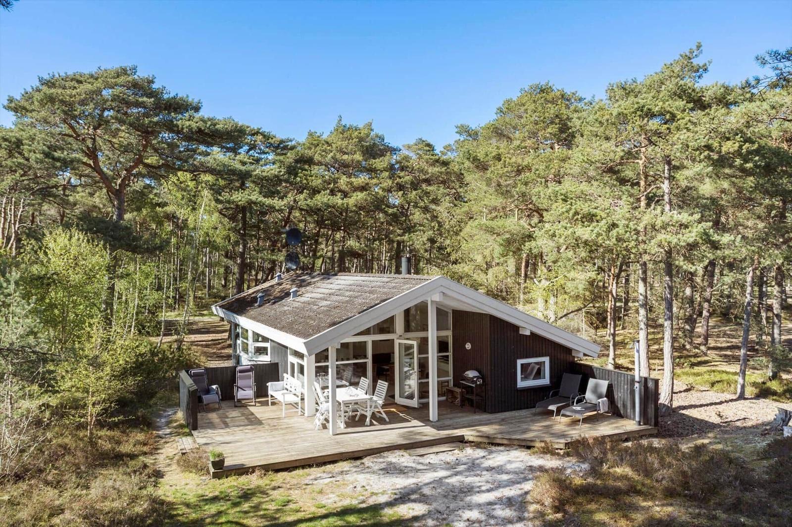 Moderne Hütte mit Holzterrasse und Grill im Wald. Weiße Möbel und große Fenster.