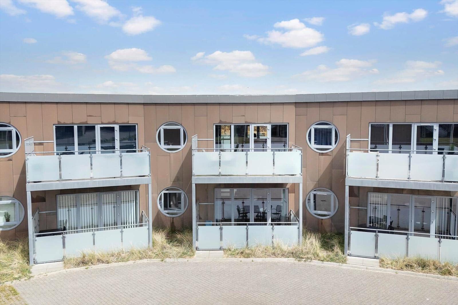 Moderne Ferienwohnungen mit Balkonen und runden Fenstern. Außenbereich mit Pflanzen und gepflastertem Platz.