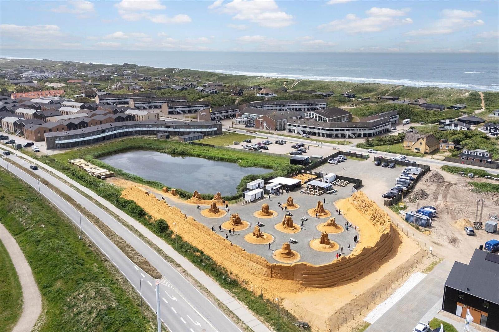Strandkunstwerk mit Sandskulpturen am Meer. Parkplatz und Gebäude im Hintergrund.