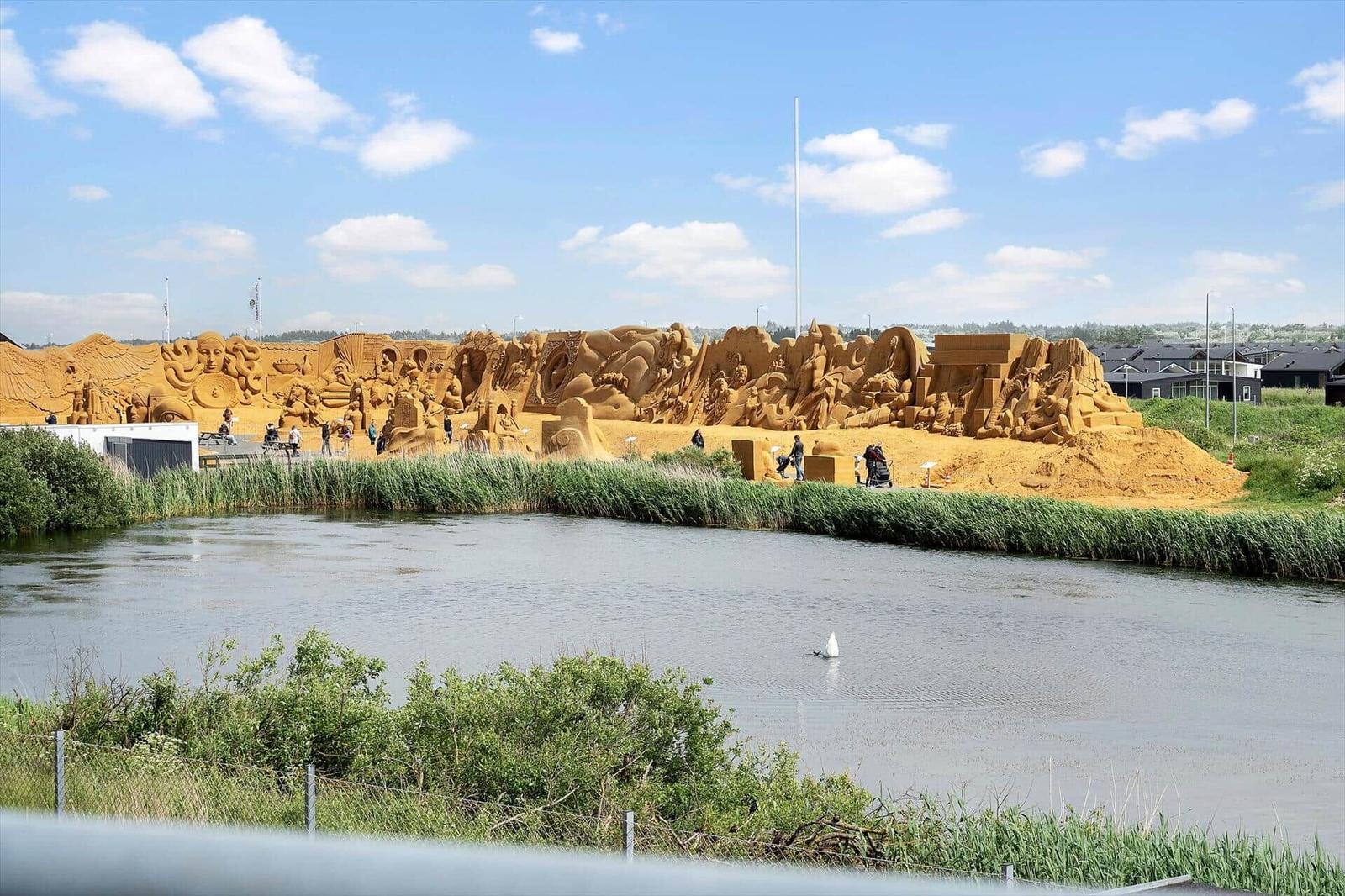 Große Sandkunst an einem See mit Besuchern und Grünflächen im Hintergrund.