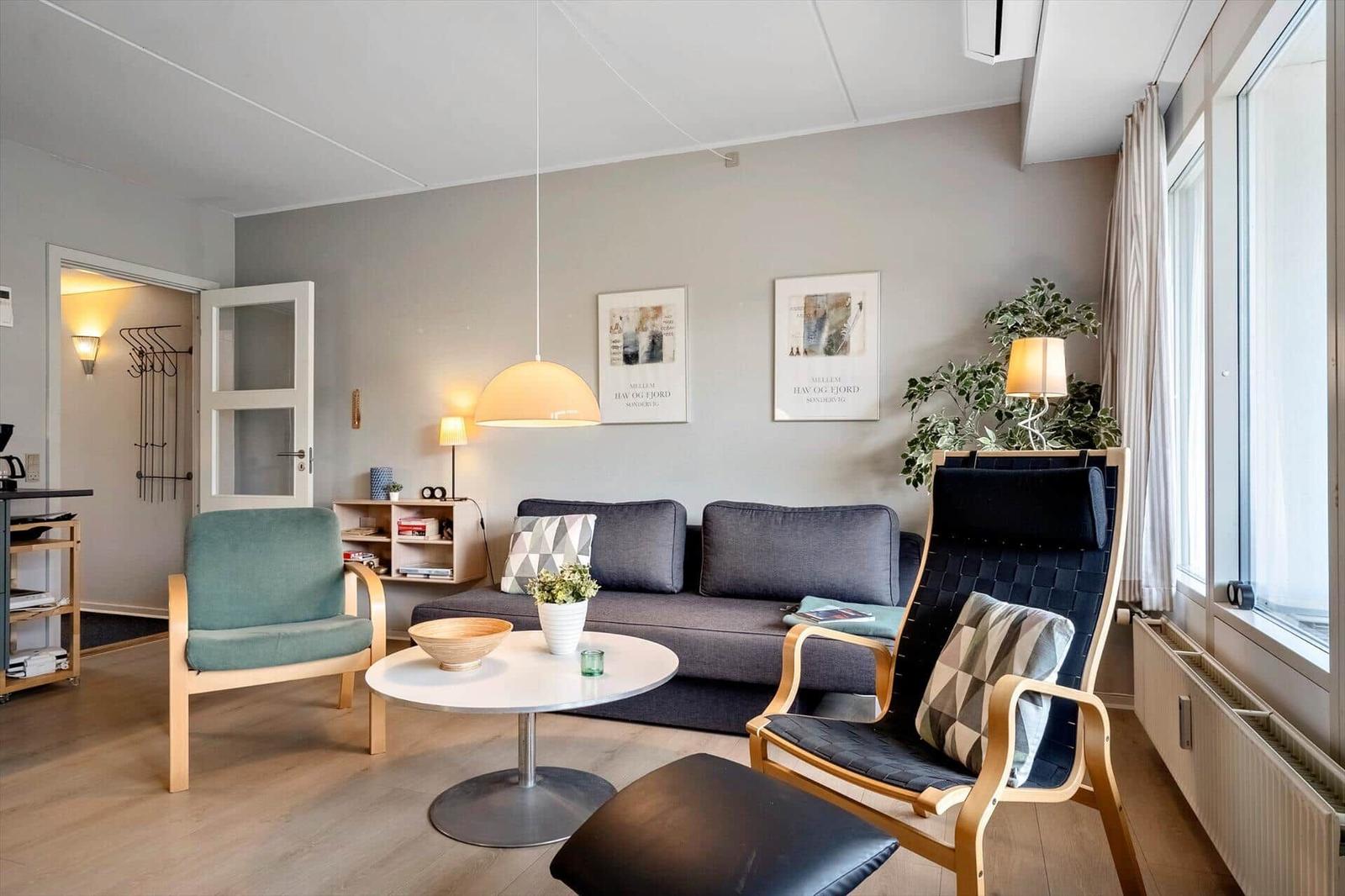 Wohnzimmer mit Sofa, zwei Stühlen und Tisch. Fenster, Pflanzen und Wandbilder.