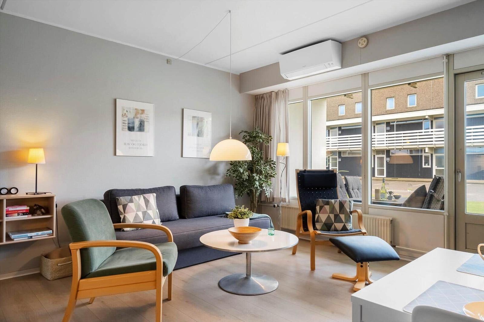 Wohnzimmer mit Sofa, Sessel und Tisch. Große Fensterfront mit Blick auf Gebäude.